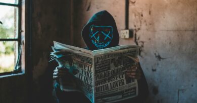 A person in a neon mask and hoodie reads a newspaper in an eerie, abandoned room.