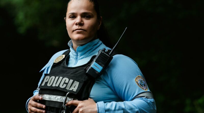 Female police officer in uniform standing outdoors with a confident expression and radio handset.