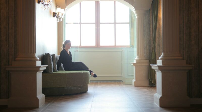 A woman in a black dress sits elegantly in a classic lobby framed by architectural columns.