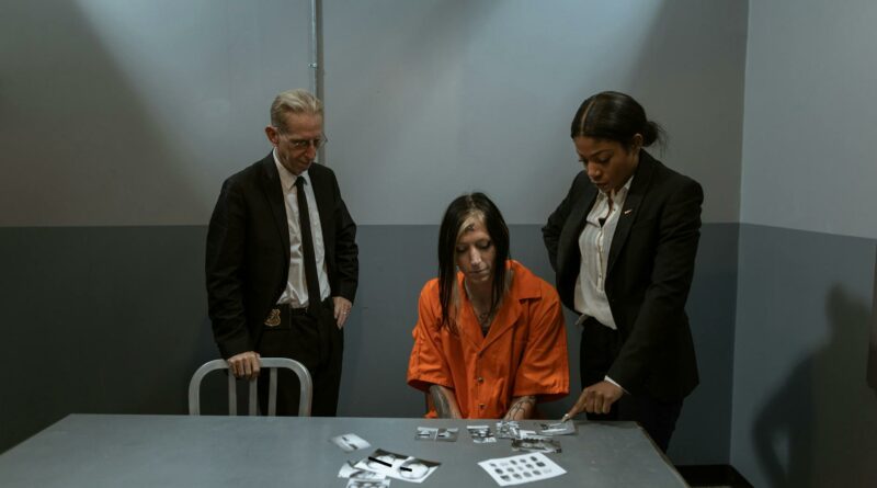 A suspect in an orange jumpsuit is questioned by detectives in an interrogation room.