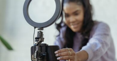 Cheerful young African American female blogger in stylish sweater smiling while setting up camera of smartphone attached to tripod with ring light before recording vlog