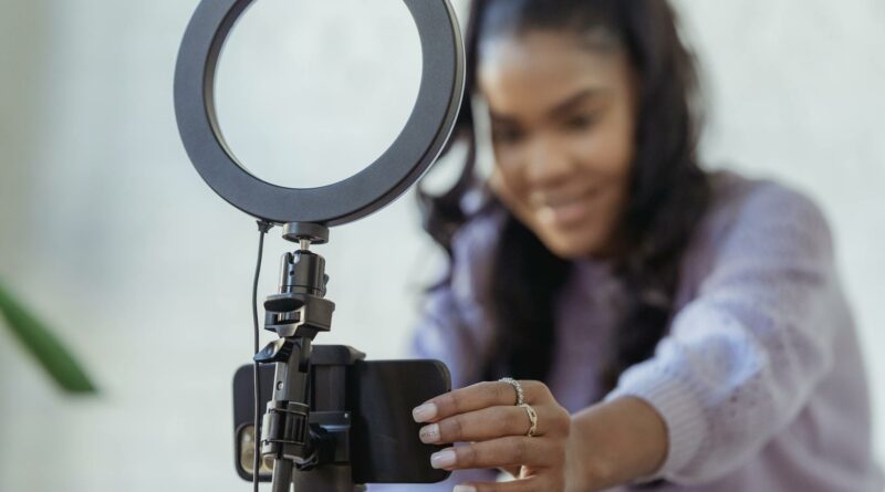Cheerful young African American female blogger in stylish sweater smiling while setting up camera of smartphone attached to tripod with ring light before recording vlog