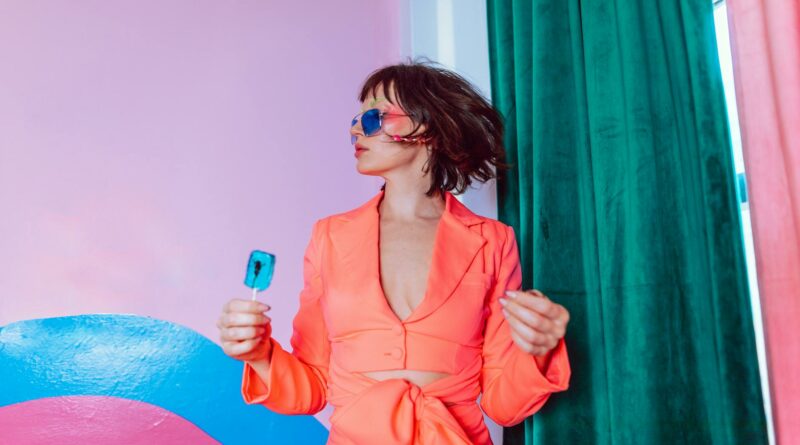 Trendy portrait featuring a woman in vibrant attire with sunglasses holding a blue lollipop.