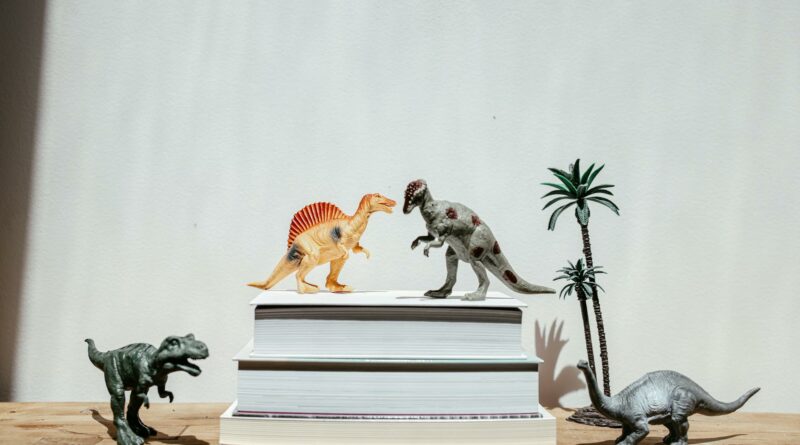 Vibrant dinosaur toys arranged on stacks of books against a plain background, creating a playful scene.