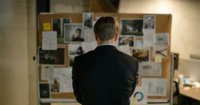 A detective in a suit examines a bulletin board with investigation materials in an office setting.