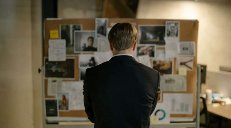 A detective in a suit examines a bulletin board with investigation materials in an office setting.