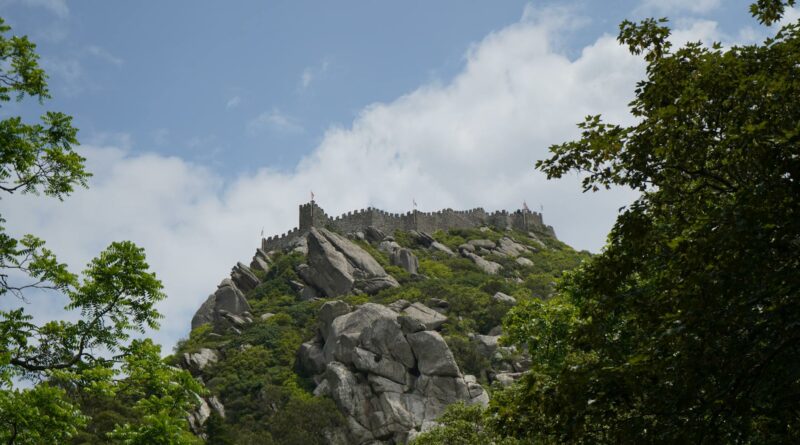 A captivating view of Castelo dos Mouros perched on a hill in Sintra, surrounded by lush greenery under a cloudy sky.