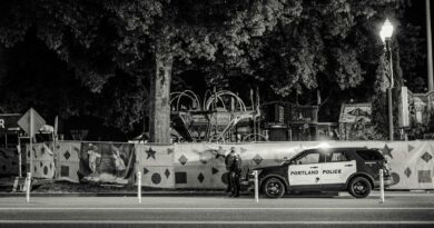 Black and white nighttime street view featuring a Portland police car parked by a decorated fence.