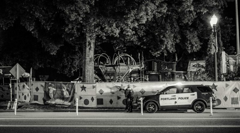 Black and white nighttime street view featuring a Portland police car parked by a decorated fence.