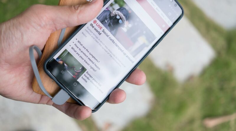 A hand holds a smartphone displaying online news while charging with a portable power bank.