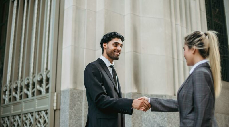 Two business professionals in formal attire shaking hands outside an elegant building, signifying partnership.