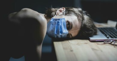 Woman wearing a face mask resting her head on a table beside a laptop, conveying pandemic fatigue and protection.