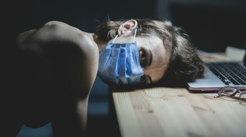 Woman wearing a face mask resting her head on a table beside a laptop, conveying pandemic fatigue and protection.