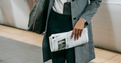 From above of crop anonymous female employee in elegant outfit and name tag standing on platform of metro station with newspaper in hand