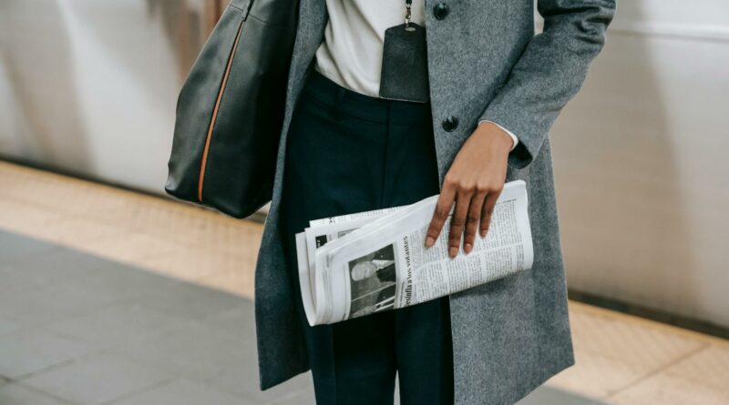 From above of crop anonymous female employee in elegant outfit and name tag standing on platform of metro station with newspaper in hand