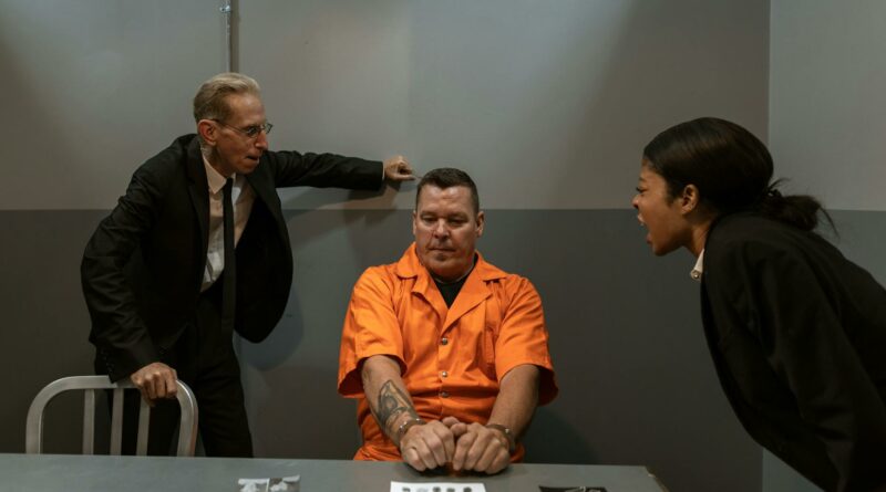 An intense interrogation with two detectives and a handcuffed prisoner in an orange jumpsuit.