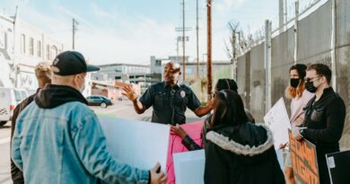 A police officer addresses a group of peaceful protesters holding signs on a city street under a clear sky.