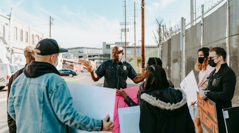A police officer addresses a group of peaceful protesters holding signs on a city street under a clear sky.