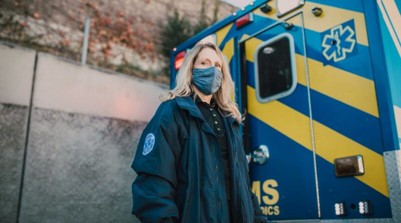 Female paramedic in a mask standing confidently by an EMS ambulance.