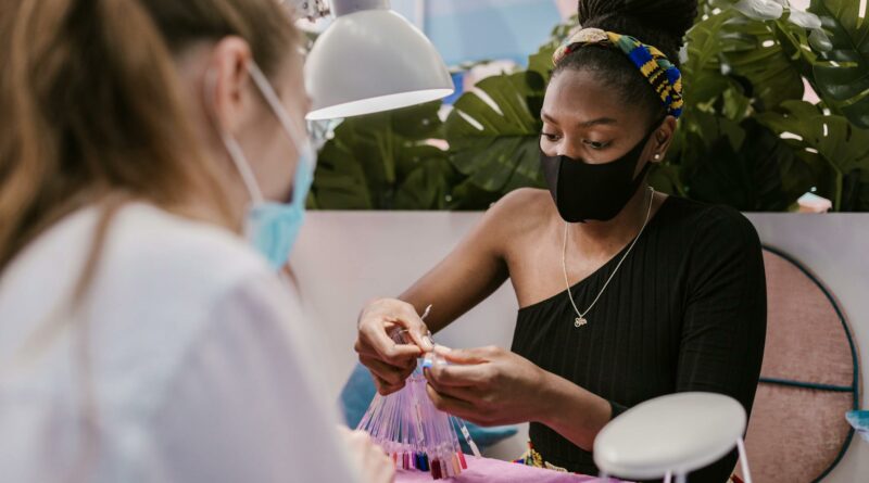 A nail technician and customer wearing masks in a modern salon choosing nail polish colors, embracing safety measures.