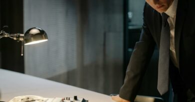 A detective in a suit examines evidence on a table under a lamp, focused on solving a case.