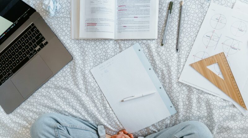 Flat lay of a study area with an open laptop, books, notes, and stationery on a bed.