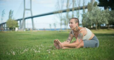 Tattooed woman in activewear stretching in park by bridge, promoting fitness and wellness.