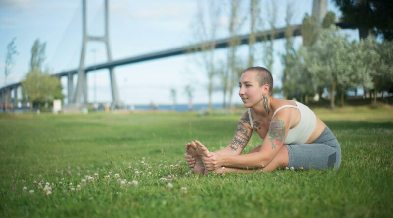 Tattooed woman in activewear stretching in park by bridge, promoting fitness and wellness.