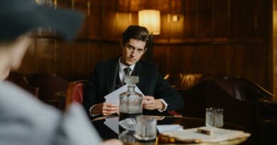 A serious man in a suit sitting at a table with papers, evoking a corporate or mobster vibe in an elegant, dimly lit room.