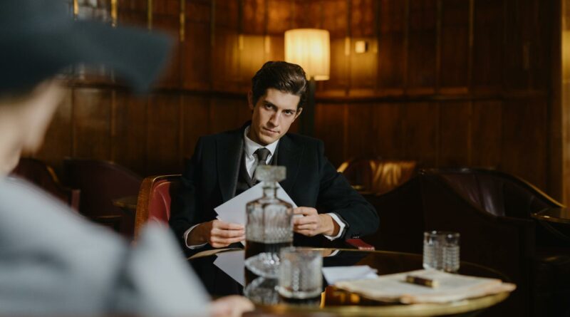 A serious man in a suit sitting at a table with papers, evoking a corporate or mobster vibe in an elegant, dimly lit room.