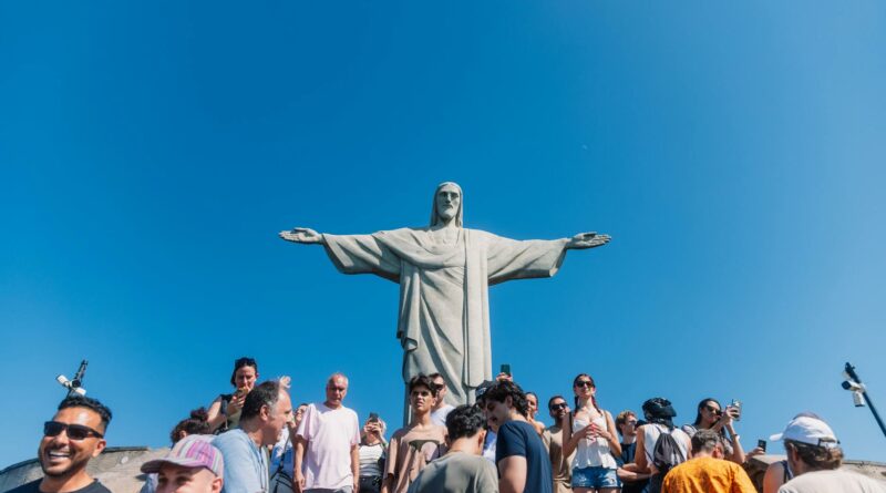 Tourists gather at the iconic Christ the Redeemer statue under clear blue skies in Rio de Janeiro, Brazil.