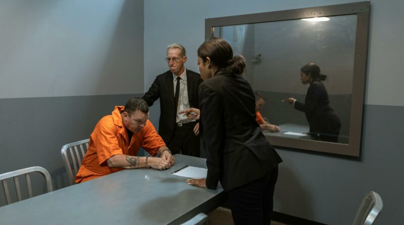 A tense interrogation scene showing a prisoner, lawyer, and detective in a police station room.