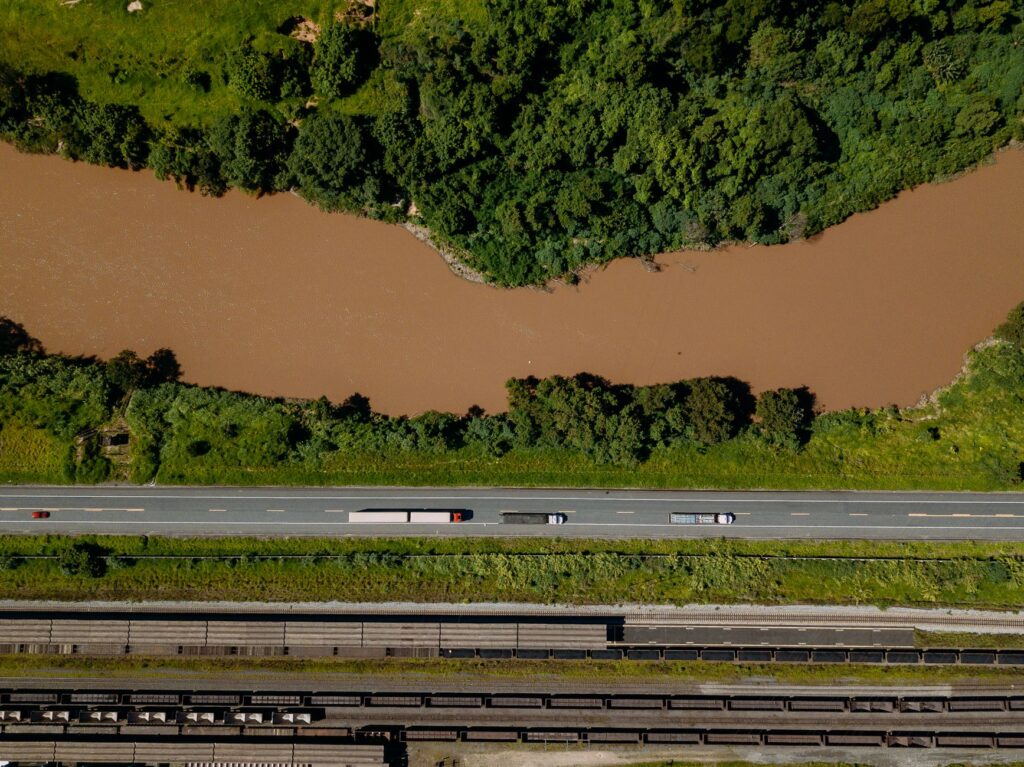 Vista aérea de uma rodovia movimentada, um trem de carga em trilhos e um porto com navios, simbolizando a infraestrutura logística do Brasil.