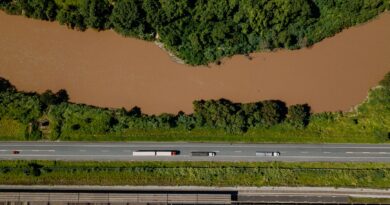 Vista aérea de uma rodovia movimentada, um trem de carga em trilhos e um porto com navios, simbolizando a infraestrutura logística do Brasil.