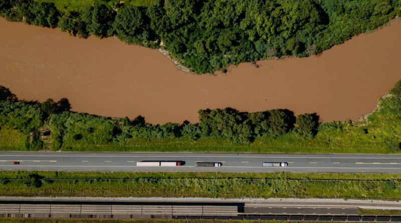 Vista aérea de uma rodovia movimentada, um trem de carga em trilhos e um porto com navios, simbolizando a infraestrutura logística do Brasil.