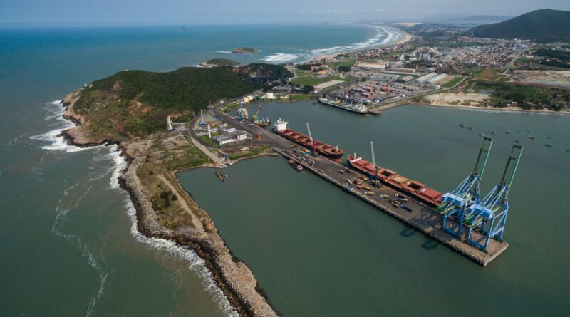 Vista aérea de uma rodovia movimentada, um trem de carga passando e navios em um porto, simbolizando a infraestrutura logística do Brasil.